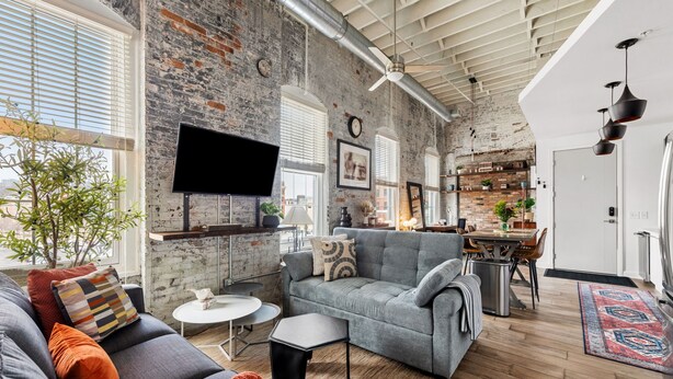 A spacious living area with a gray sofa, a small round table, and a television mounted on a brick wall.