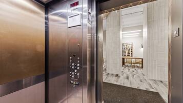 Um elevador com painel de controle e vista para um saguão com mesa e cadeiras.