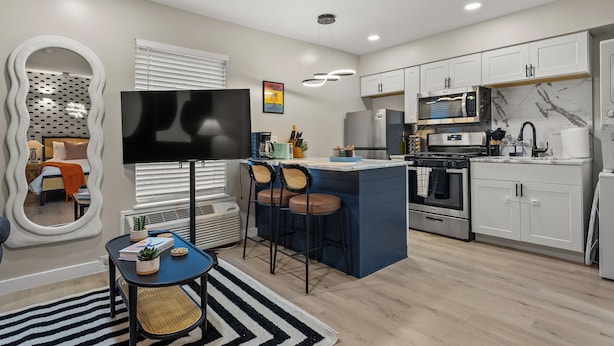 A modern kitchen with white cabinets, a built-in microwave, and a stove. There is a bar area with stools and a flat-screen TV. - Cozy Queen Bed + Sofa Bed Studio | Free Parking | Mins to DT | VAMC | UTHSC (Memphis)