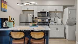 A modern kitchen with white cabinets, a stainless steel microwave, a built-in oven, and a washing machine. There are two chairs with woven backs and brown cushions at the island. - Cozy Queen Bed + Sofa Bed Studio | Free Parking | Mins to DT | VAMC | UTHSC (Memphis)