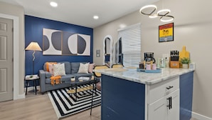 A modern kitchen with a central island, a dining area with a sofa and coffee table, and a living room with a blue accent wall and abstract art. - Cozy Queen Bed + Sofa Bed Studio | Free Parking | Mins to DT | VAMC | UTHSC (Memphis)