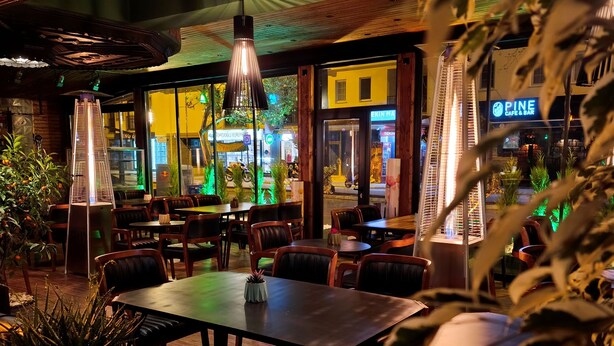 Un área de comedor al aire libre con mesas y sillas, rodeada de plantas y vista a un paisaje urbano nocturno.