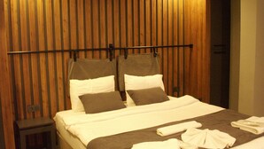 Habitación de hotel con cabecera de madera, cama con ropa de cama blanca y gris, y una mesita de noche pequeña.