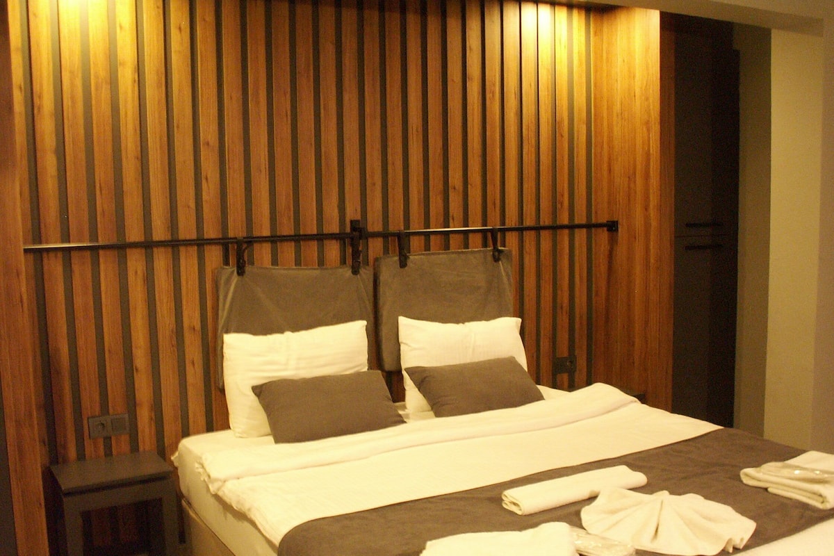 A hotel room with a wooden slat headboard, a bed with white and gray bedding, and a small bedside table.