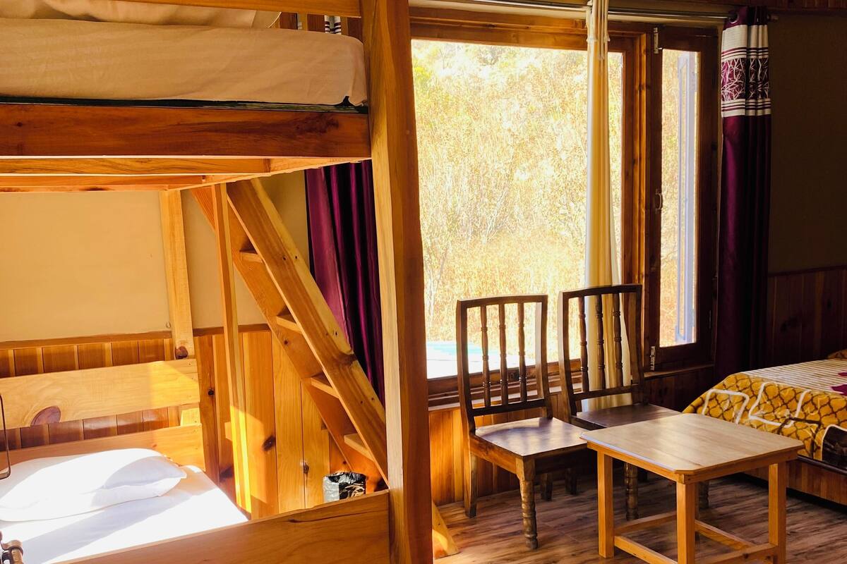 A bunk bed with a wooden ladder, a wooden table, and chairs in a room with wooden flooring and a window with curtains.