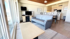 Ein modernes Wohnzimmer mit einer Couch, einem Couchtisch und einem Fernseher. Es gibt eine Küchenzeile mit einem Esstisch und Stühlen.