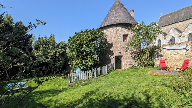 Un edificio de piedra con techo cónico, una puerta de madera y una ventana pequeña. Hay una cerca de piquetes blancos y un área verde con dos sillas rojas.