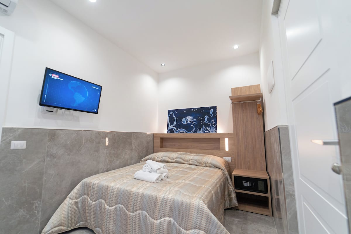 A modern hotel room with a bed, a flat-screen TV mounted on the wall, a wooden headboard, and a built-in wardrobe.