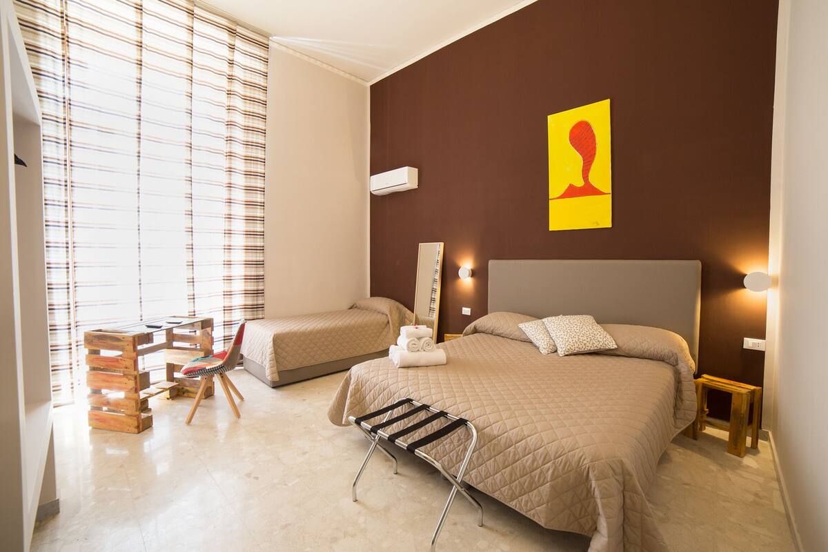 A hotel room with two beds, a wooden desk, a chair, and a window with blinds.