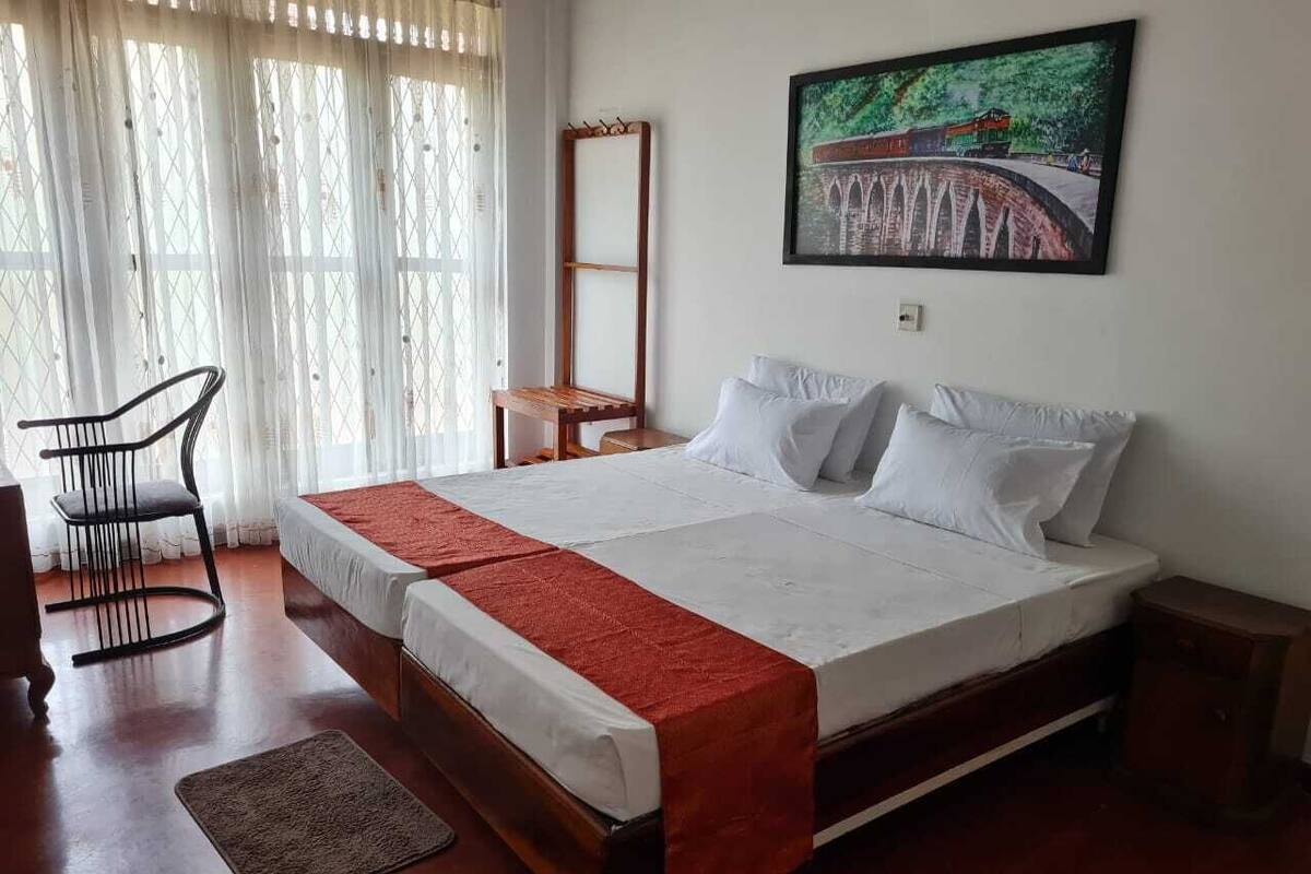 A double bed with a red runner, a wooden nightstand, a chair, and a painting on the wall.