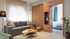 A modern hotel room with a grey sofa, a round wooden coffee table, a green armchair, a wooden cabinet, and a potted plant.