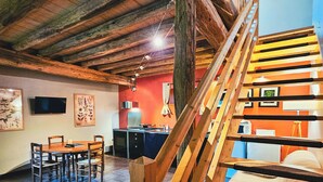 A rustic interior with a wooden staircase, a dining area with chairs and a table, and a kitchen in the background. - 1BDR Apt + Sofa Bed w/ Garden + Jacuzzi (Étivey)