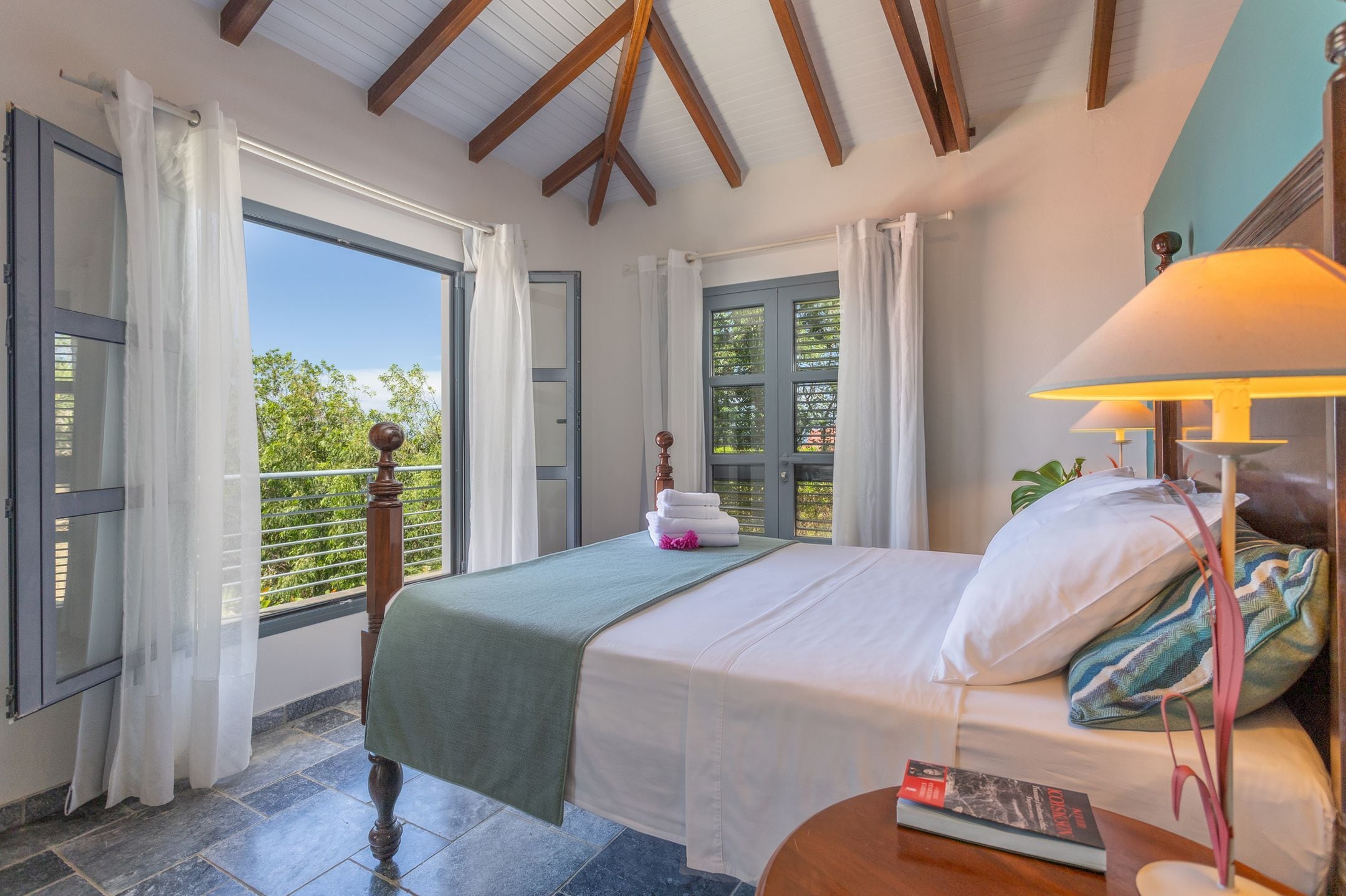 A bedroom with a bed, a bedside table, a lamp, and a view of greenery outside.