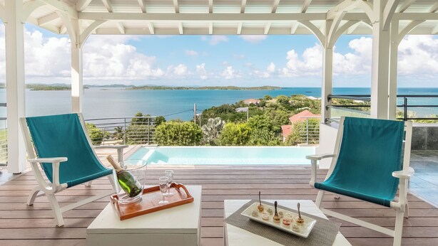 Ein überdachter Außenbereich mit zwei Stühlen und einem Tisch, mit Blick auf einen Pool und eine malerische Aussicht auf das Meer und das entfernte Land.
