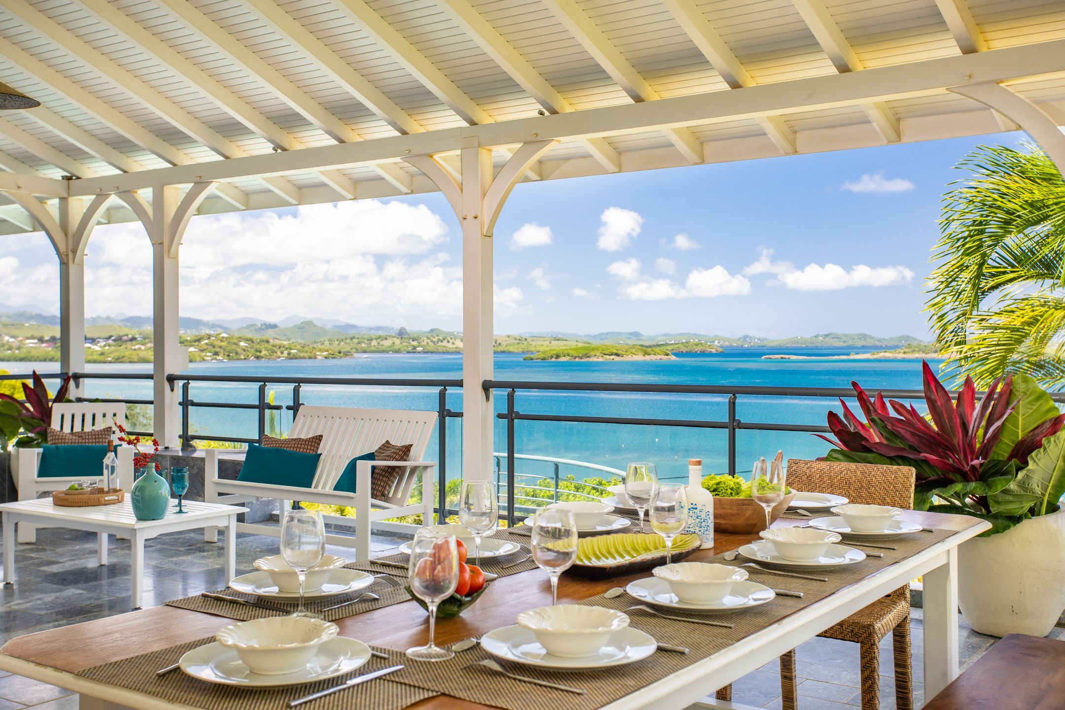 Un área de comedor al aire libre con vista al mar y a las colinas, amueblada con mesas y sillas blancas, y una estructura de techo cubierta.