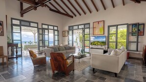 Ein geräumiges Wohnzimmer mit hoher Decke, großen Fenstern und Meerblick. Zwei Sofas, ein Holzstuhl und ein Couchtisch sind vorhanden.