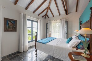 A bedroom with a bed, curtains, a window, a painting, and a nightstand with a lamp. - Villa Big Blue  (Le François)