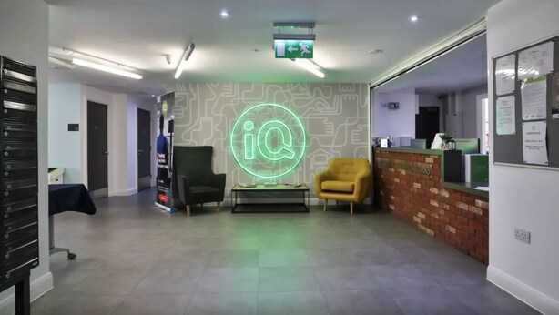 Un espace d'accueil moderne avec un panneau néon représentant le logo « iQ », un comptoir d'accueil, un fauteuil jaune et un mur décoratif en briques.
