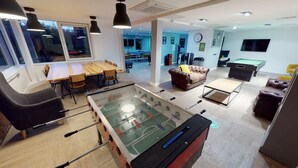 Il s'agit d'un espace intérieur moderne comprenant un baby-foot, une table de billard et une table de flipper. On y trouve un coin repas avec une table et des chaises en bois, ainsi qu'un canapé. La pièce est dotée de grandes fenêtres et d'une horloge murale.