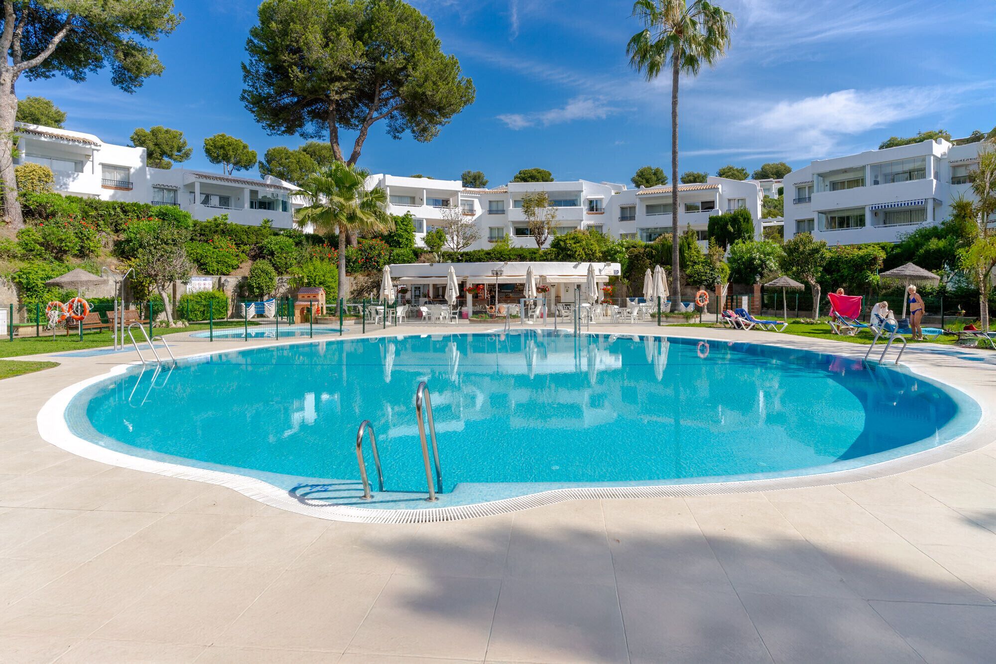 A large swimming pool surrounded by white buildings, with clear blue skies and greenery.
