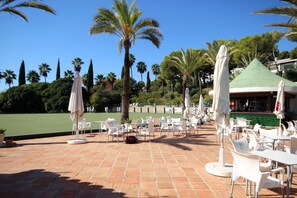 An outdoor seating area with white chairs and umbrellas, palm trees, and a green tennis court in the background. - Spacious studio apartment in Miraflores (Mijas Costa)
