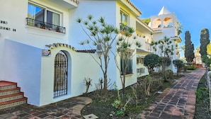 A building with a white facade, a tiled roof, and a small garden with various plants. - Spacious studio apartment in Miraflores (Mijas Costa)