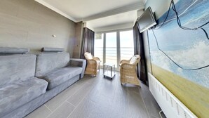 A modern living room with a grey sofa, wicker chairs, a television mounted on the wall, and a large window offering a view of the sea.