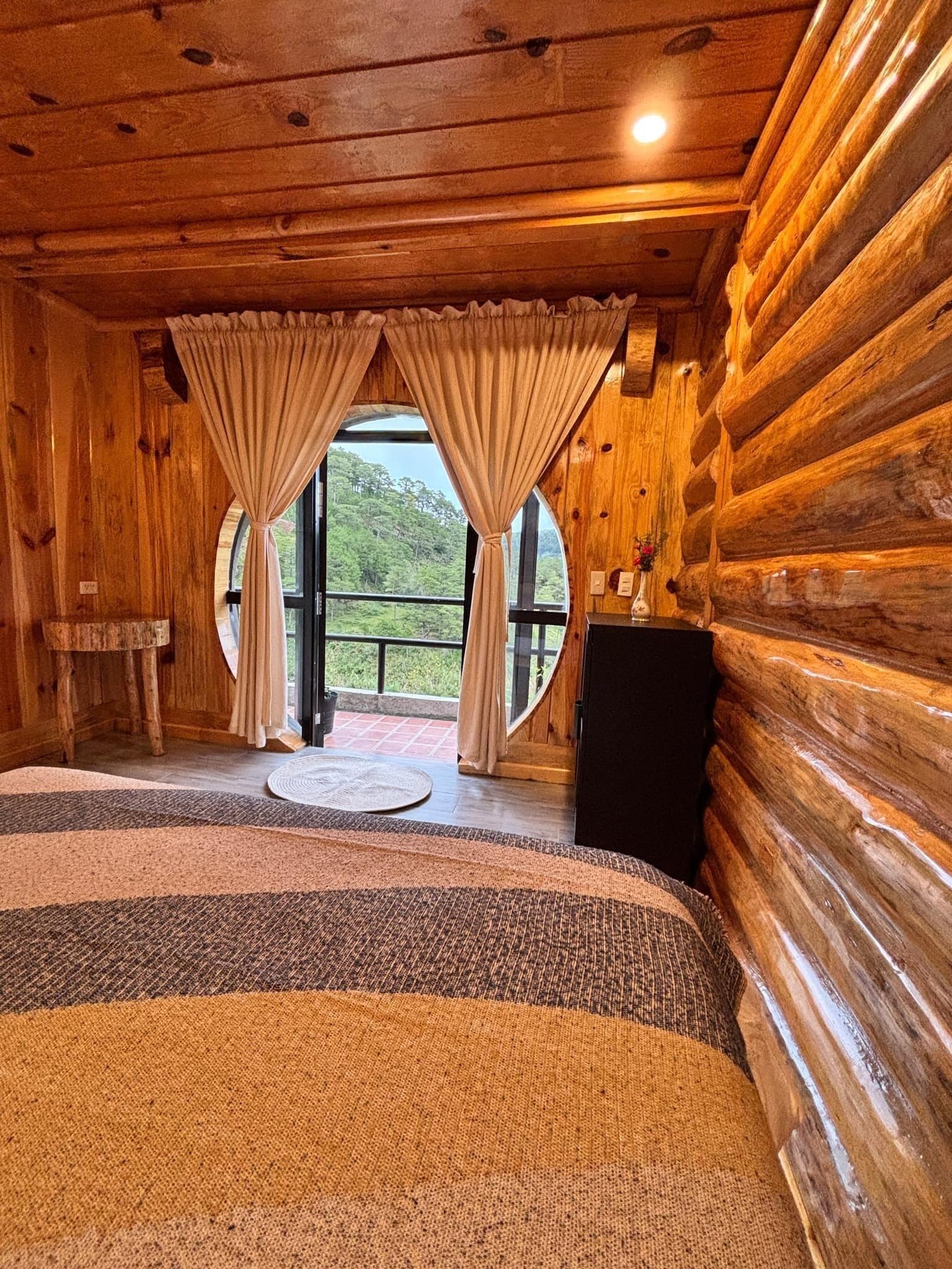 Ein Schlafzimmer in einer Blockhütte mit einem großen Bett, zwei mit Vorhängen versehenen Fenstern, einem kleinen Tisch und Blick auf die grüne Landschaft.