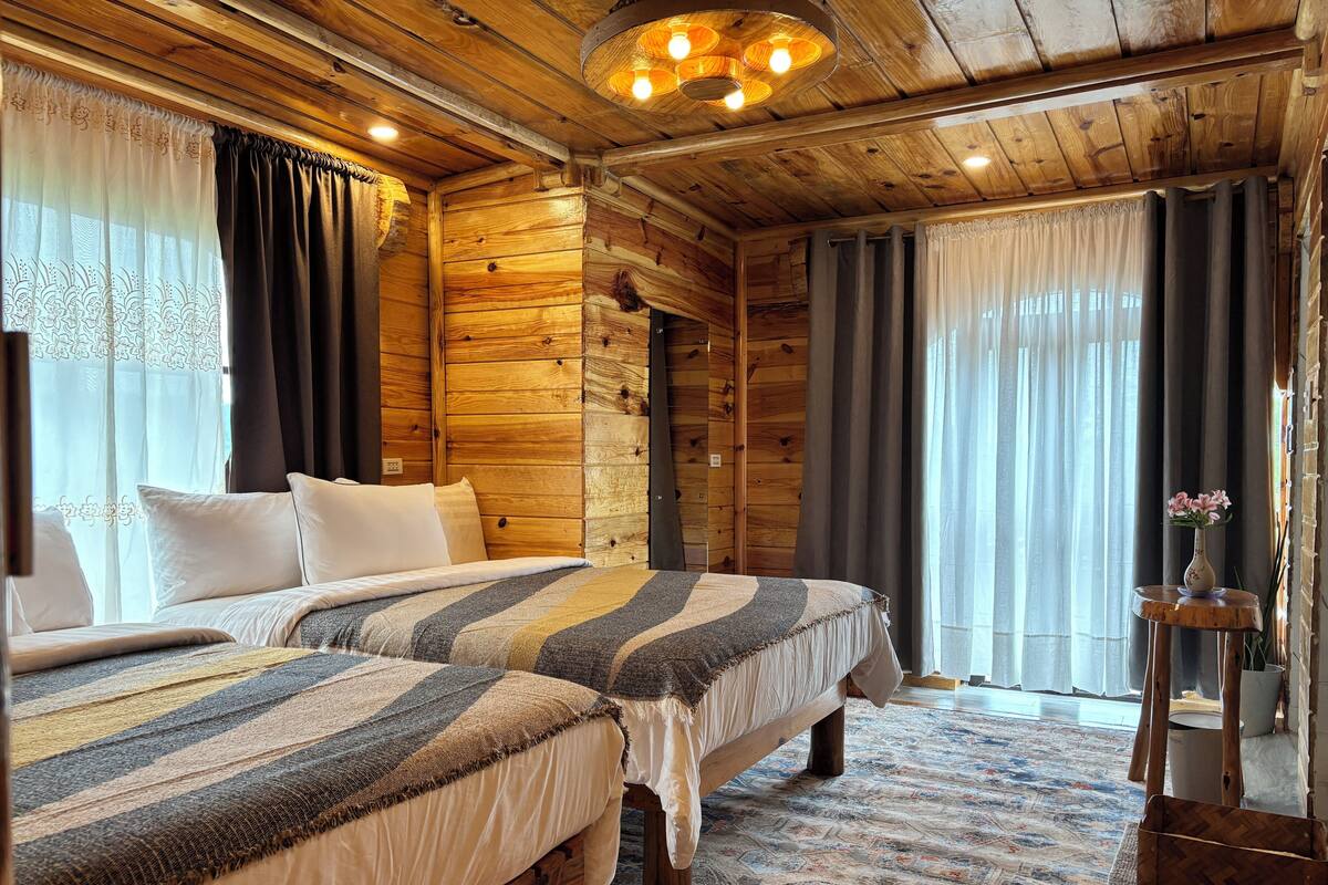 A cozy log cabin bedroom with two beds, a wooden ceiling, and a large window with curtains.