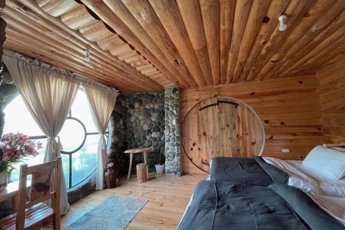 A cozy cabin interior with a wooden ceiling, a stone wall, a round wooden door, a bed, a wooden table with chairs, and a window with curtains.
