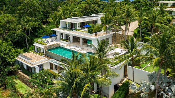 A modern multi-story house with a swimming pool, surrounded by lush greenery and palm trees.