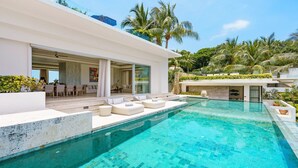 A modern poolside area with a clear blue pool, outdoor seating, and lush greenery in the background.