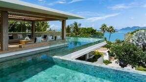 Una piscina infinita moderna con borde de cristal, una terraza de madera con asientos y vistas al océano y las montañas.