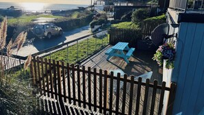 Une terrasse avec une table de pique-nique et une jardinière suspendue, d’où l’on a vue sur une route et une zone côtière.