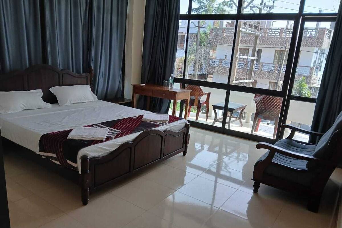 A hotel room with a large bed, a small table, two chairs, and a view of the outside through large windows.