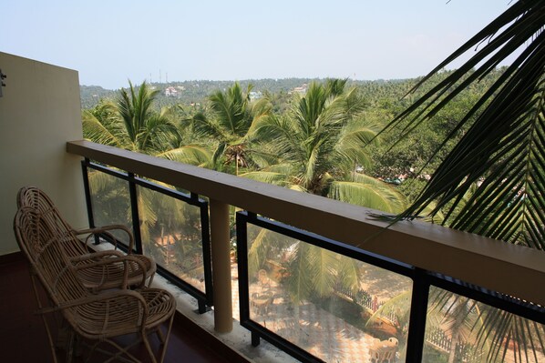A balcony with wicker chairs, a glass railing, and a view of palm trees and buildings in the distance.