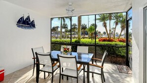 Une terrasse ensoleillée avec une table et des chaises en verre, un ventilateur de plafond et une vue sur des palmiers et un parking.