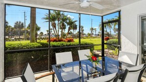 Une véranda avec une table et des chaises en verre, un ventilateur de plafond et une vue sur un jardin aménagé, agrémenté de palmiers et de fleurs.