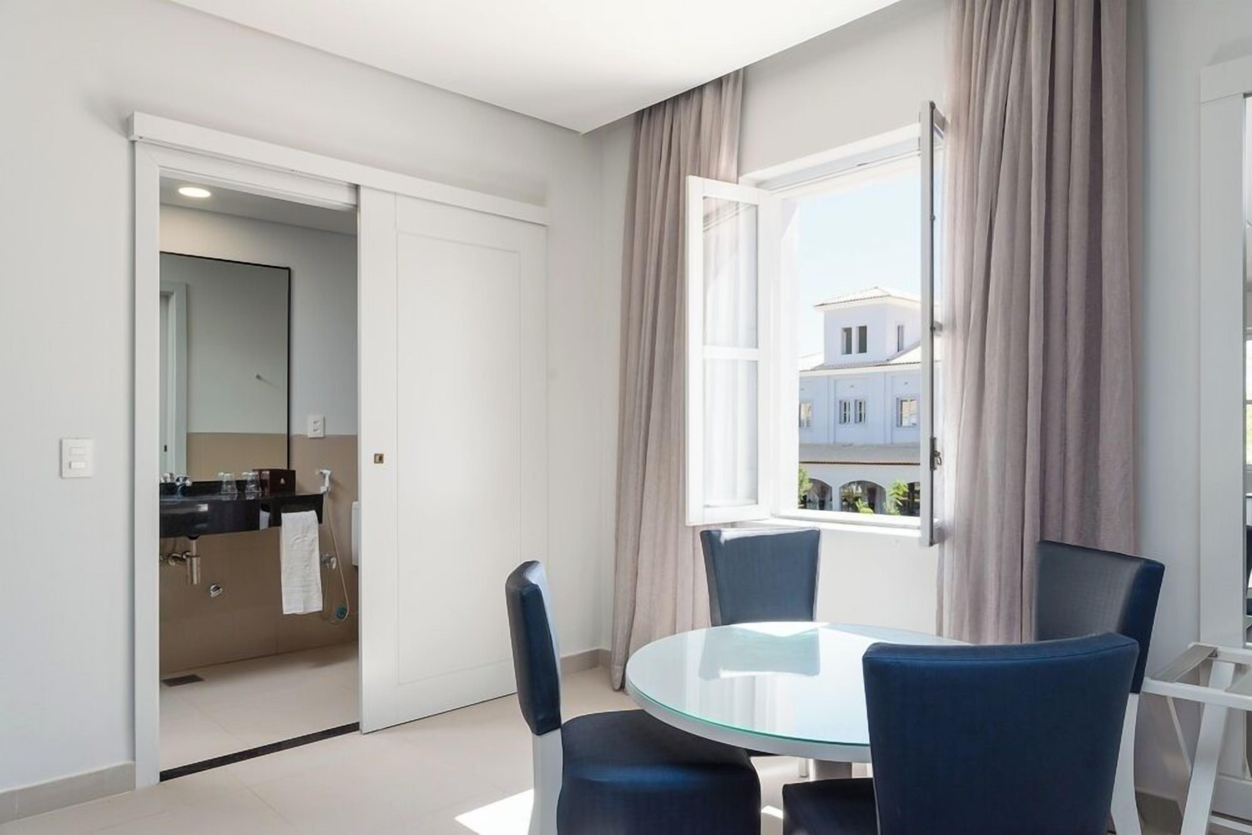 A modern room with a glass table and four chairs, a window with curtains, and a view of buildings outside.