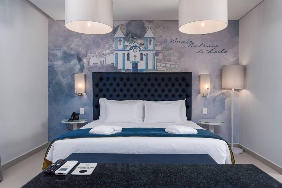 Hotelkamer met een groot bed, een hoofdeinde met een stadsgezicht, twee bedlampen, een tafeltje met een telefoon en afstandsbedieningen, en een kunstwerk aan de muur.