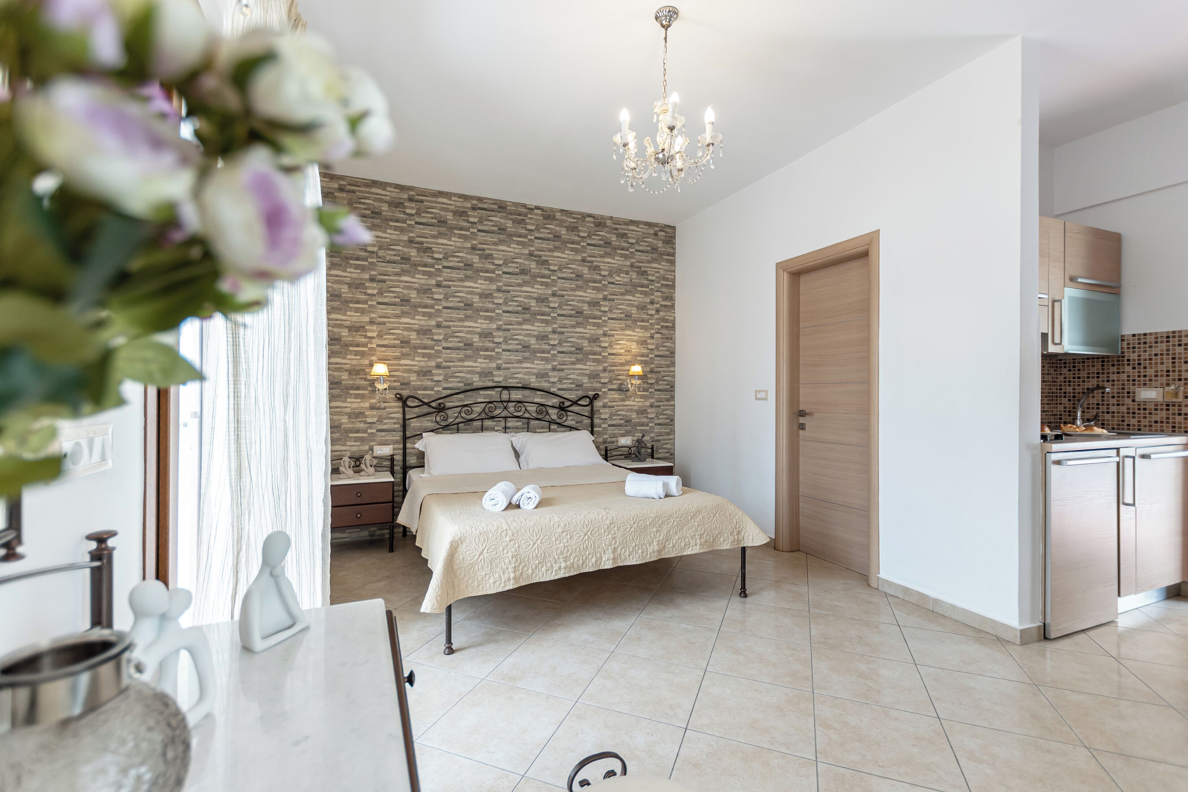 A modern bedroom with a bed, bedside tables, a chandelier, and a view of the kitchen.