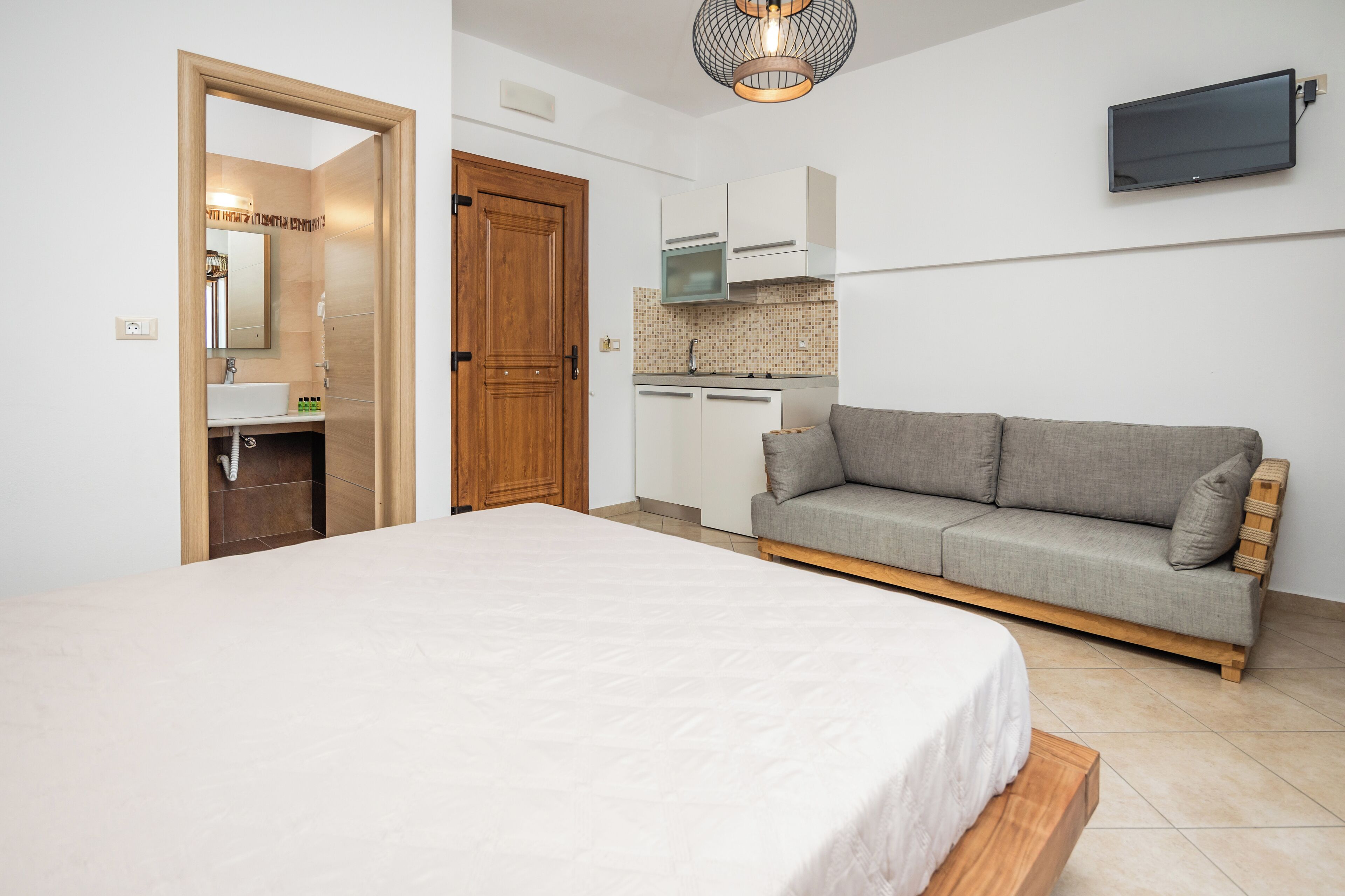 A bedroom with a bed, a sofa, a kitchen area, and a TV mounted on the wall.