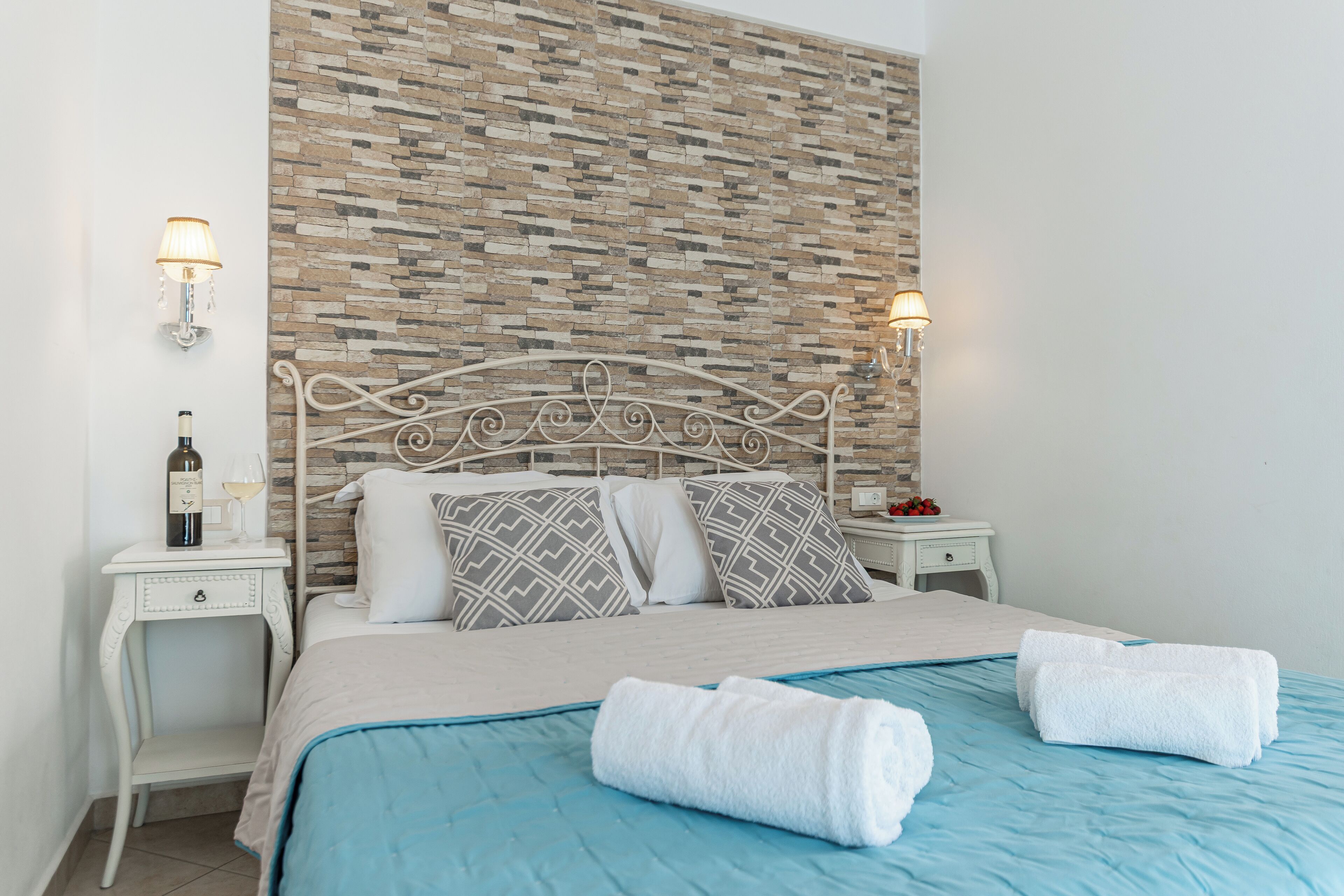 Ein ordentlich bezogenes Bett mit blauer Bettdecke und zwei aufgerollten Handtüchern, ein weißer Nachttisch mit Flasche und Glas sowie eine Wand mit einer strukturierten Steinoptik.