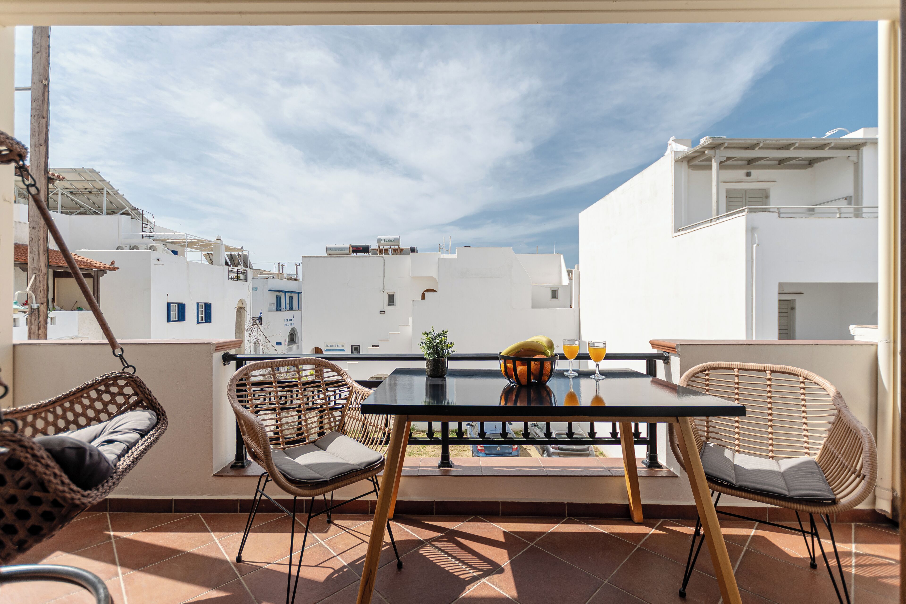 A balcony with wicker chairs, a table, and a view of white buildings under a clear sky.