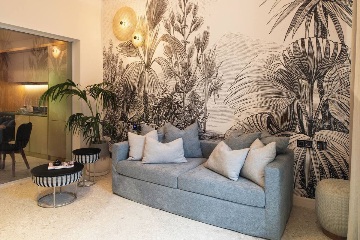 Una sala de estar con un sofá gris, dos mesitas redondas y un gran mural en la pared con plantas tropicales.