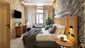 Une chambre à coucher avec un lit, une télévision, une fenêtre avec des rideaux, une plante et un mur orné d’un motif de feuilles.