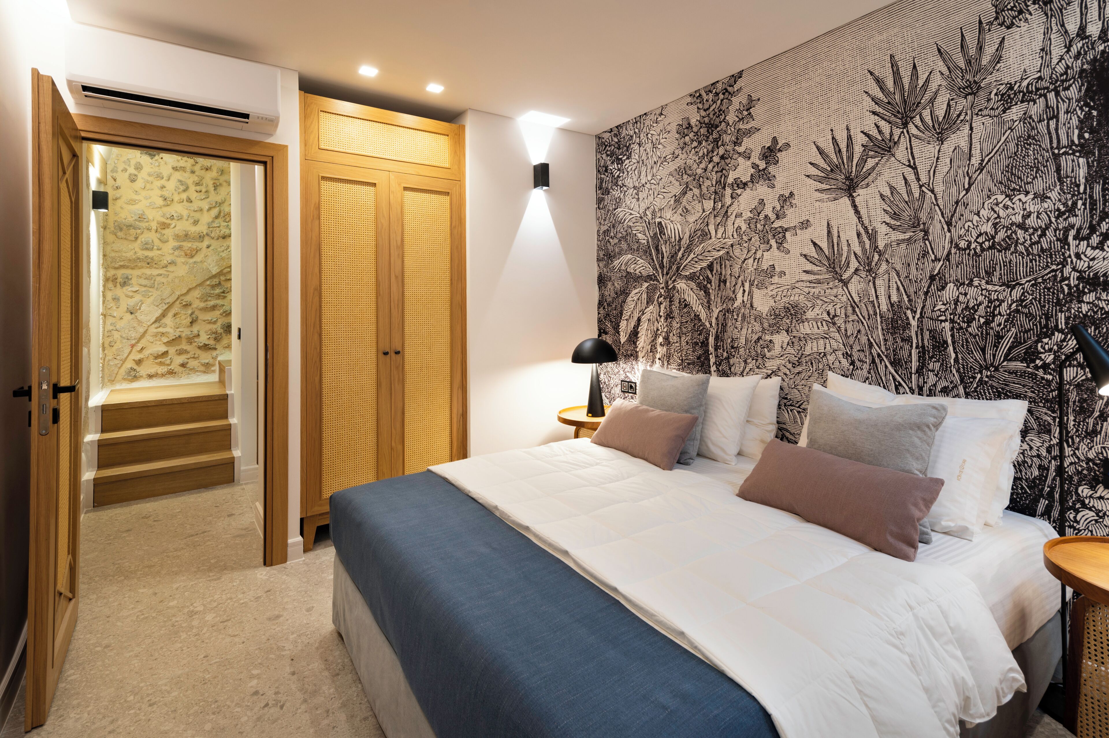 A bedroom with a large bed, a nightstand, a wall-mounted lamp, and a patterned wallpaper.