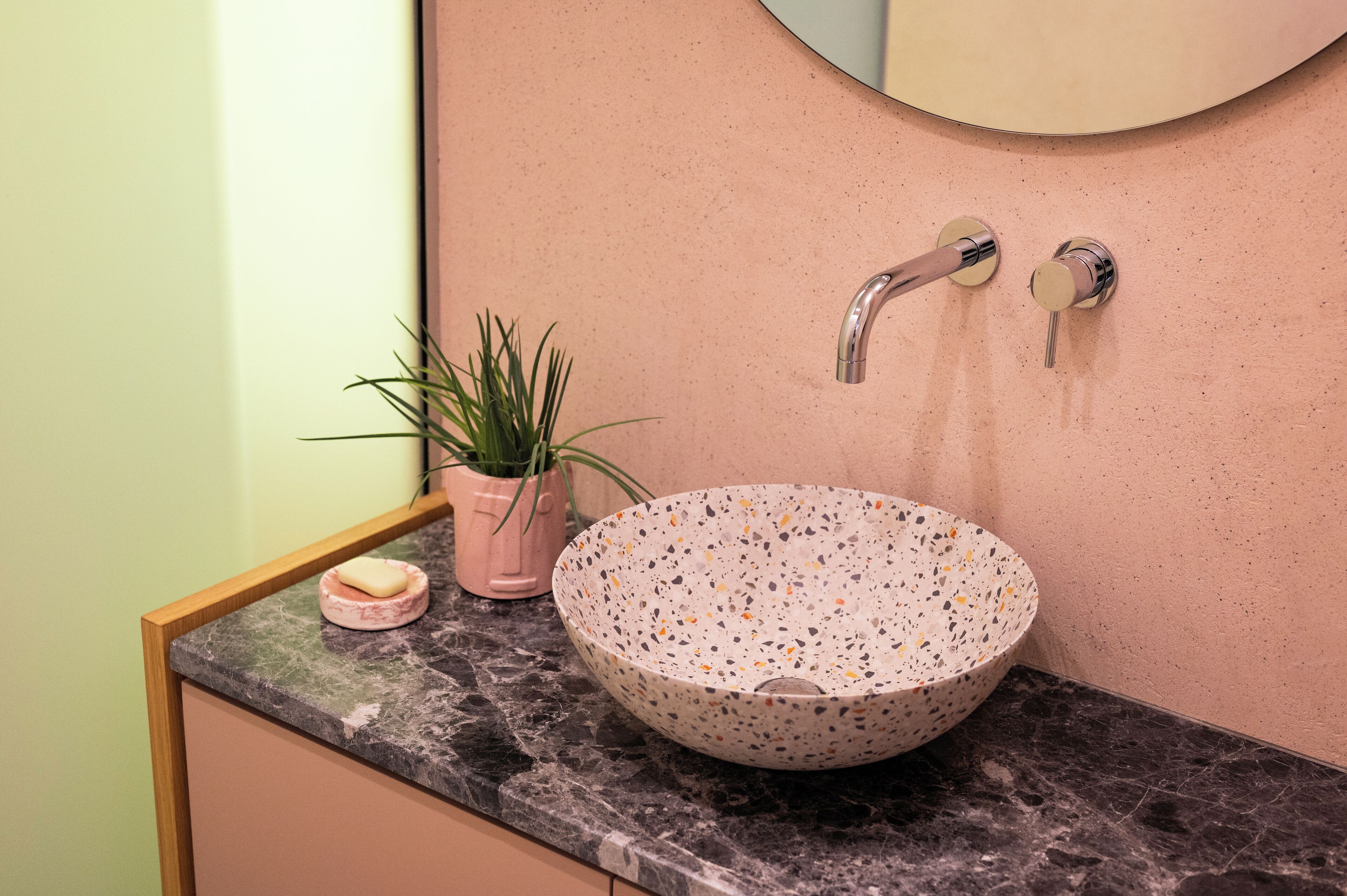 Un baño con un lavamanos redondo y moteado, una planta rosa y un espejo con marco de madera.