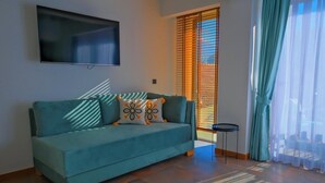 A teal sofa with patterned cushions, a mounted TV, a small side table, and a view through a window with blinds.