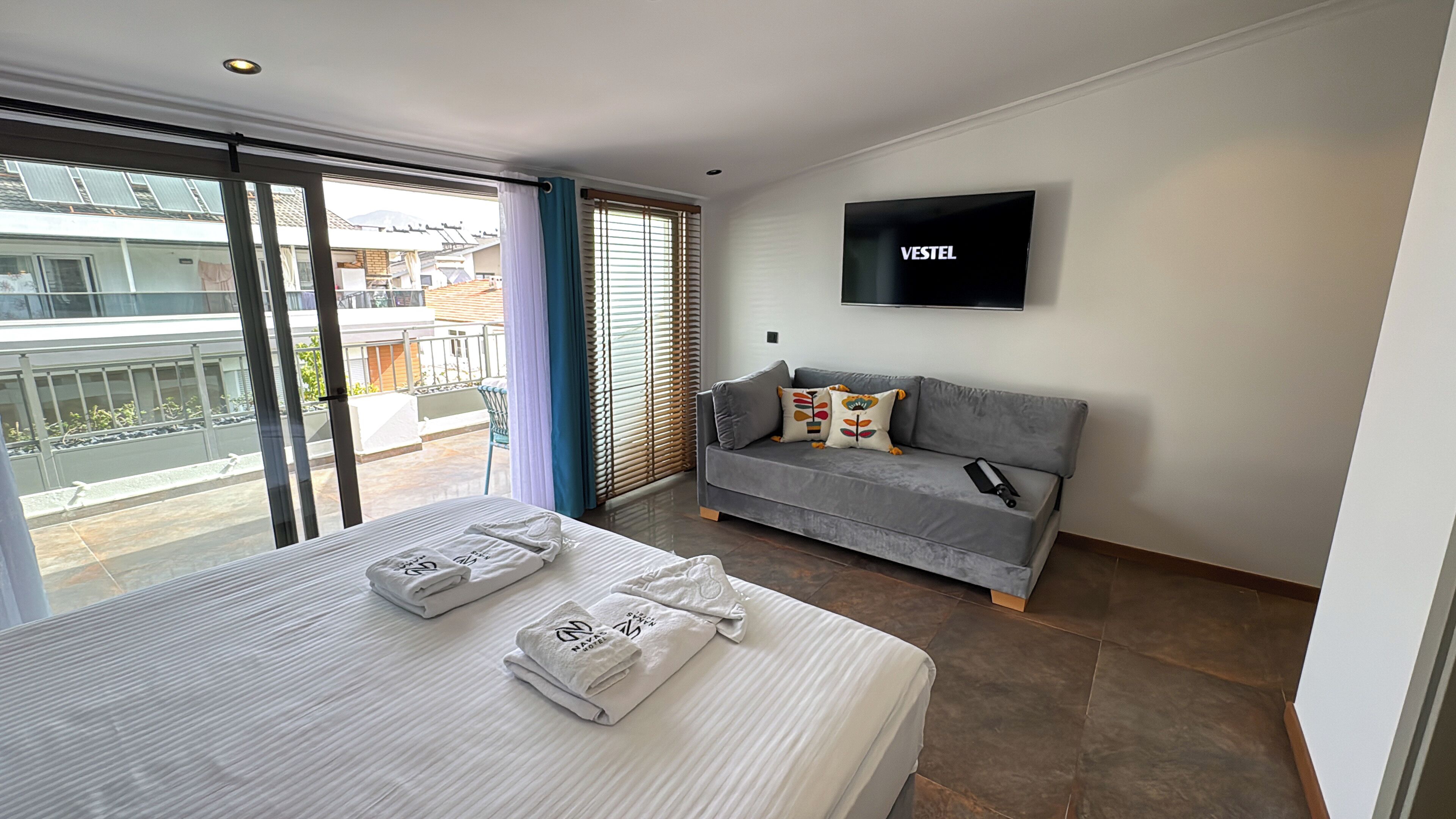 A hotel room with a bed, a grey sofa, a TV, and a balcony with a view of buildings.
